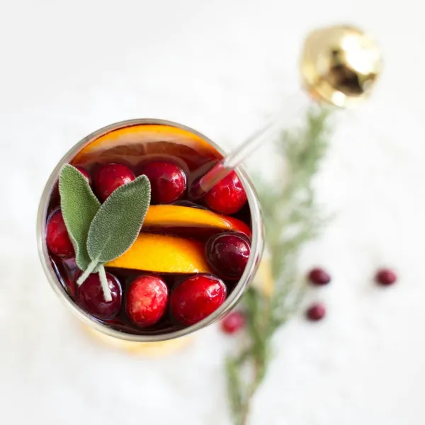 Un verre de cocktail rempli de canneberges rouges, de tranches d'orange, de feuilles de sauge, avec une garniture de romarin à côté.