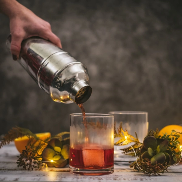 Un mixologue verse un cocktail rouge dans un verre à whisky rempli de glaçons, entouré de tranches d'orange et de décorations végétales sur une table en bois.
