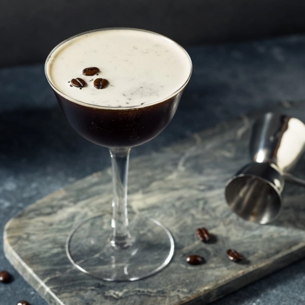 Un verre à cocktail élégant rempli d'un Espresso Martini crémeux, décoré de trois grains de café sur le dessus, posé sur une surface en marbre gris avec un doseur métallique à côté.
