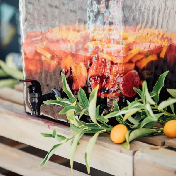 Un grand distributeur en verre rempli de sangria rouge avec des tranches d'orange, de citron et de fruits rouges visibles, entouré de branches décoratives vertes et de petites oranges sur une surface en bois.