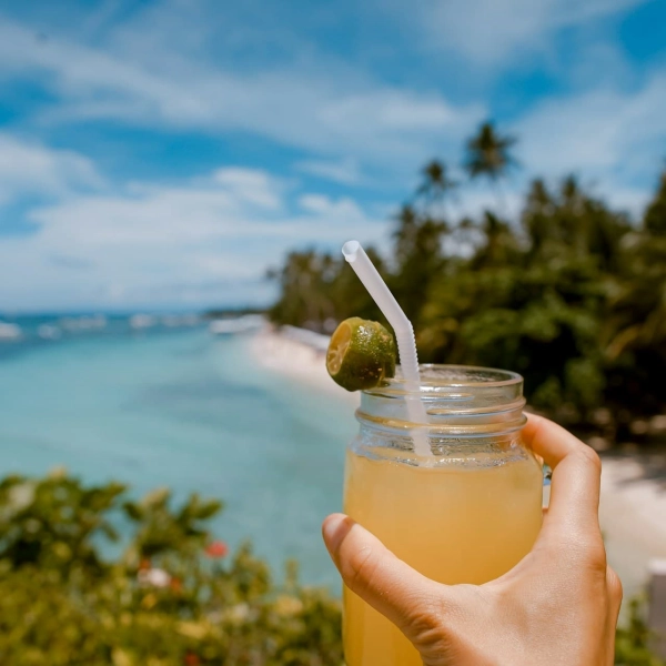 Un verre de punch au rhum garni d'une tranche de citron vert, tenu par une main au bord d'une plage tropicale avec une mer turquoise et un ciel bleu en arrière-plan.