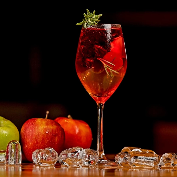 Verre à cocktail rempli de boisson rouge garnie de romarin frais, entouré de glaçons transparents et de pommes rouges et vertes.