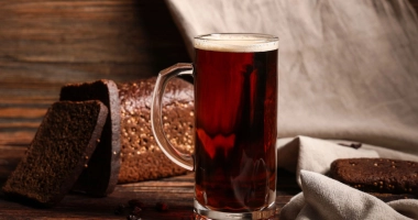 Une chope de bière brunâtre à côté d'une miche de pain, posée sur une table en bois avec une toile de fond de tissu beige.