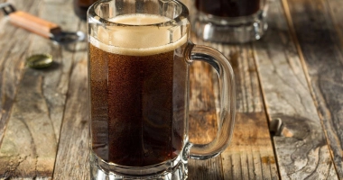 Un grand verre à cocktail avec du soda brun foncé, une mousse crémeuse débordante sur le dessus, décoré d'une paille rayée verte et blanche, marqué du logo "BEST DAMN" à l'avant.