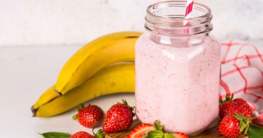 Milkshake à la fraise et banane, servi dans un verre à pied, garni d'une tranche de fraise sur le bord, entouré de fraises fraîches et de bananes.