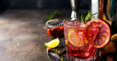 Verre de cocktail rempli de Greyhound rose pâle, garni d'un brin de romarin, avec un demi-pamplemousse flou en arrière-plan.