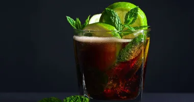 Un verre de Cuba Libre avec glaçons, décoré de quartiers de citron vert, sur une table en bois avec deux morceaux de citron vert à côté.