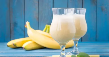 Deux verres remplis de banana daiquiri glacé, garnis de tranches de banane et de citron vert, avec une feuille de menthe sur le dessus, posés sur un dessous de verre à motifs bleus et blancs.