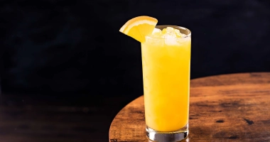Verre long rempli de cocktail jaune vif garni d'une tranche d'orange, entouré de tranches d'orange fraîche.