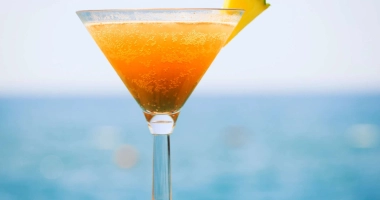 Verre de cocktail sur la plage contenant un mélange de liquide jaune et rouge, garni d'une tranche d'ananas, d'une cerise, d'un petit parasol rose et d'une paille rouge.