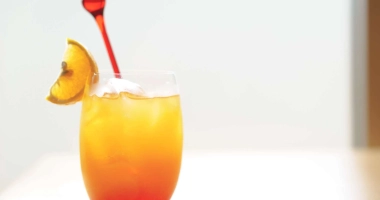 Verre de cocktail avec dégradé de couleurs rouge en bas et jaune en haut, garni d'une tranche d'orange et d'une cerise rouge.