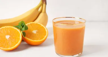 Verre de cocktail apello avec un jus orange clair, entouré de fruits tropicaux comme bananes et ananas en arrière-plan.