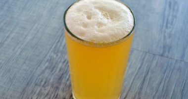 Verre à pied rempli d'un cocktail mousseux jaune pâle sur une table en bois, éclairé par la lumière.