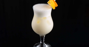 Un verre de margarita blanche garni de noix de coco râpée, avec une tranche d'orange et une cerise rouge en décoration sur le bord du verre.