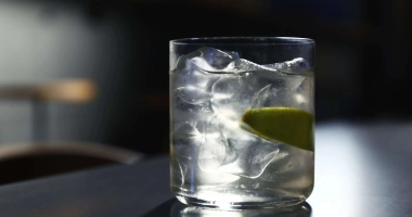 Verre de cocktail transparent rempli de glaçons, avec un quartier de citron et une cerise sur un bâtonnet, surmonté d'une boisson claire versée.