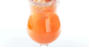 Un cocktail rose pâle servi dans un verre rempli de glaçons, garni d'une tranche d'ananas et d'une cerise, avec une paille bleue.