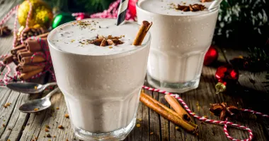 Verres de cocktail remplis de lait de poule crémeux, garnis de bâtons de cannelle et saupoudrés de muscade, posés sur une table en bois chaleureuse.