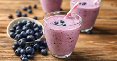 Verre de smoothie rose crémeux avec des myrtilles fraîches sur une brochette décorative, posé sur une nappe bleue.