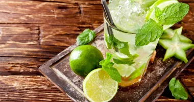 Verre de Mojito alsacien avec de la menthe fraîche, tranches de citron vert, et glaçons, posé sur une table en bois, avec une demi-lime à côté.