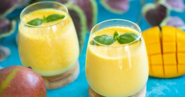 Verre de lassi à la mangue crémeux avec morceaux de mangue en décoration, posé sur une surface sombre, une mangue entière à côté.