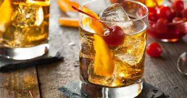 Un verre à cocktail rempli d'un mélange rouge foncé, garni d'une tranche d'orange sur le bord, avec une boisson en arrière-plan.