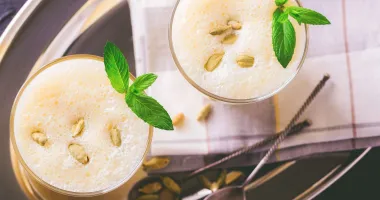 Verre de lassi blanc crémeux garni d'une feuille de menthe, avec une paille rayée sur une surface en bois.