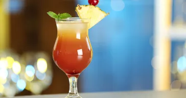 Un verre de cocktail rum runner avec une boisson rouge-orange, garni d'une brochette de fruits comprenant un morceau d'ananas et une fraise, avec une torsade de zeste de citron sur le côté.