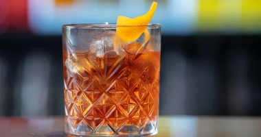 Un verre à cocktail rempli de glaçons, contenant un liquide ambré, surmonté d'une pelure d'orange et d'une griottine, avec une paille en métal.