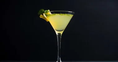 Un verre à martini rempli d'un cocktail jaune vif, garni de zestes de citron, avec un demi-citron posé à côté sur une table en bois.