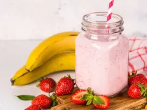 Milkshake à la fraise et banane, servi dans un verre à pied, garni d'une tranche de fraise sur le bord, entouré de fraises fraîches et de bananes.