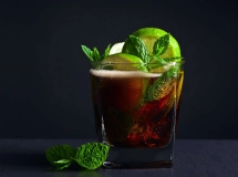 Un verre de Cuba Libre avec glaçons, décoré de quartiers de citron vert, sur une table en bois avec deux morceaux de citron vert à côté.