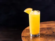 Verre long rempli de cocktail jaune vif garni d'une tranche d'orange, entouré de tranches d'orange fraîche.