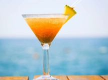 Verre de cocktail sur la plage contenant un mélange de liquide jaune et rouge, garni d'une tranche d'ananas, d'une cerise, d'un petit parasol rose et d'une paille rouge.