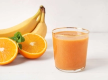 Verre de cocktail apello avec un jus orange clair, entouré de fruits tropicaux comme bananes et ananas en arrière-plan.