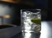 Verre de cocktail transparent rempli de glaçons, avec un quartier de citron et une cerise sur un bâtonnet, surmonté d'une boisson claire versée.