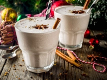 Verres de cocktail remplis de lait de poule crémeux, garnis de bâtons de cannelle et saupoudrés de muscade, posés sur une table en bois chaleureuse.