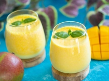 Verre de lassi à la mangue crémeux avec morceaux de mangue en décoration, posé sur une surface sombre, une mangue entière à côté.