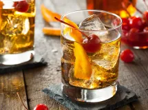 Un verre à cocktail rempli d'un mélange rouge foncé, garni d'une tranche d'orange sur le bord, avec une boisson en arrière-plan.