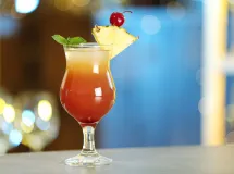 Un verre de cocktail rum runner avec une boisson rouge-orange, garni d'une brochette de fruits comprenant un morceau d'ananas et une fraise, avec une torsade de zeste de citron sur le côté.