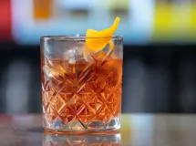 Un verre à cocktail rempli de glaçons, contenant un liquide ambré, surmonté d'une pelure d'orange et d'une griottine, avec une paille en métal.