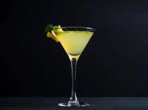 Un verre à martini rempli d'un cocktail jaune vif, garni de zestes de citron, avec un demi-citron posé à côté sur une table en bois.