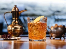 Verre à whisky rempli de cocktail ambré avec un gros glaçon, posé sur un plateau métallique.