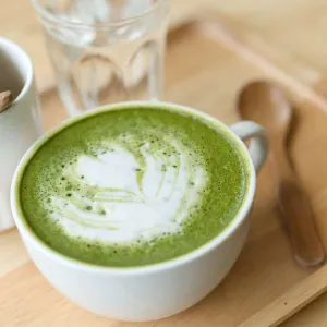 Tasse de matcha latte avec motif de feuille sur la mousse blanche, posée sur un plateau en bois avec une cuillère à côté.