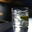 Verre de cocktail transparent rempli de glaçons, avec un quartier de citron et une cerise sur un bâtonnet, surmonté d'une boisson claire versée.