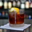 Un verre old fashioned rempli de cocktail ambré, garni d'un zeste d'orange et d'une tranche de citron, posé sur une serviette en papier blanc, avec un bar en arrière-plan flou.