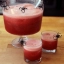 Grand bol de punch rouge mousseux avec des araignées décoratives noires sur le bord et deux verres assortis sur une table en bois.