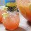 Deux verres remplis d'un cocktail rose-orange avec des glaçons et ornés d'un quartier de citron sur le bord, posés à côté d'une grande carafe contenant le même punch aux tranches de citron à l'intérieur, le tout sur une table en bois.