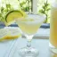 Verre à cocktail rempli d'une boisson jaune pâle avec des tranches de citron et de lime en décoration, placé sur une table avec des rondelles de citron à côté, en arrière-plan une vue floue sur un jardin.