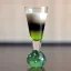 Un verre à liqueur élégant avec des couches distinctes de liquide, passant de vert vif à noir et se terminant par une mousse blanche crémeuse.