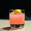 Verre à cocktail rempli d'une boisson rose vif, garnie de zestes d'agrumes jaunes flottant à la surface.